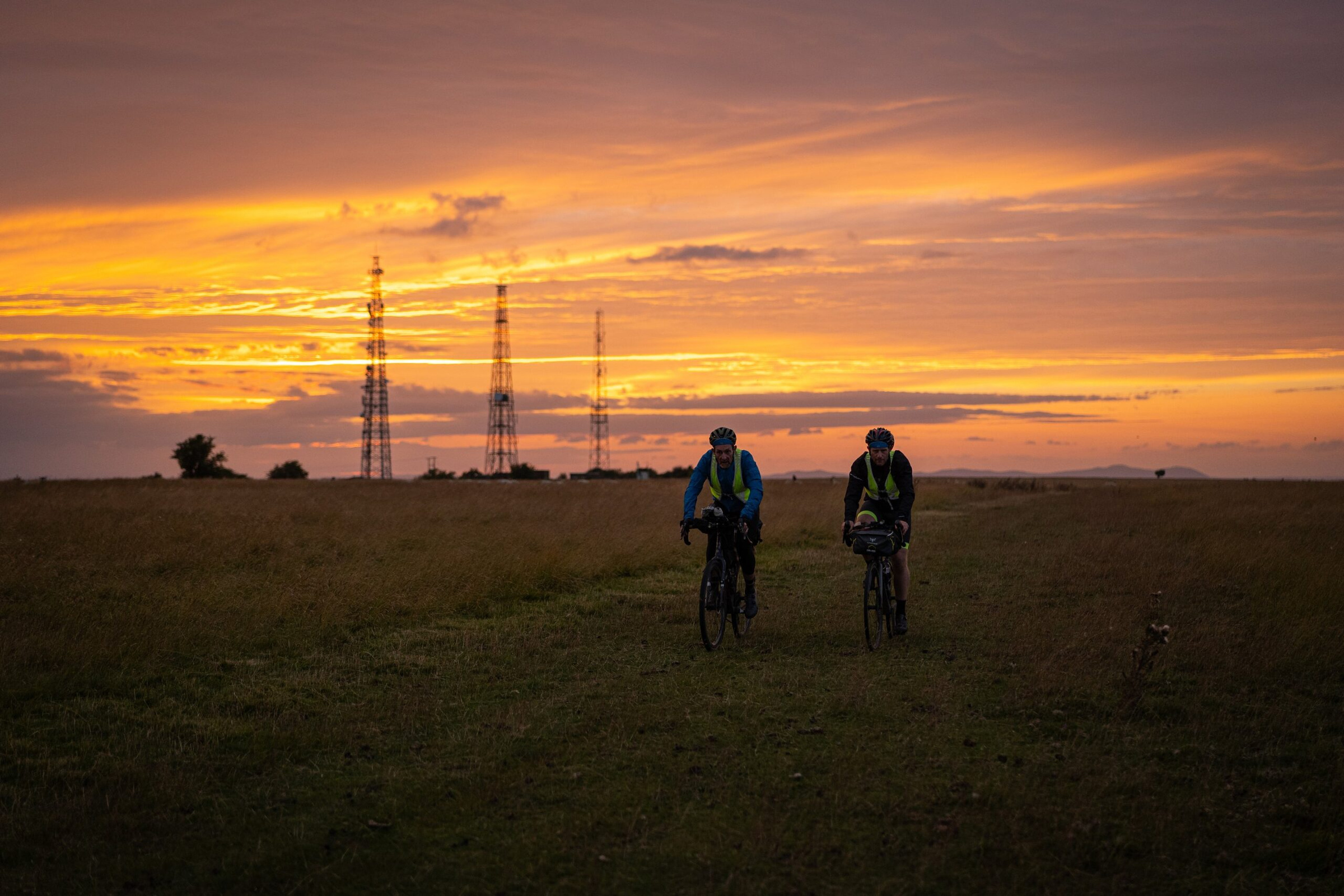 Two riders crossing Cleeve Hill at sunset beneath the radio masts