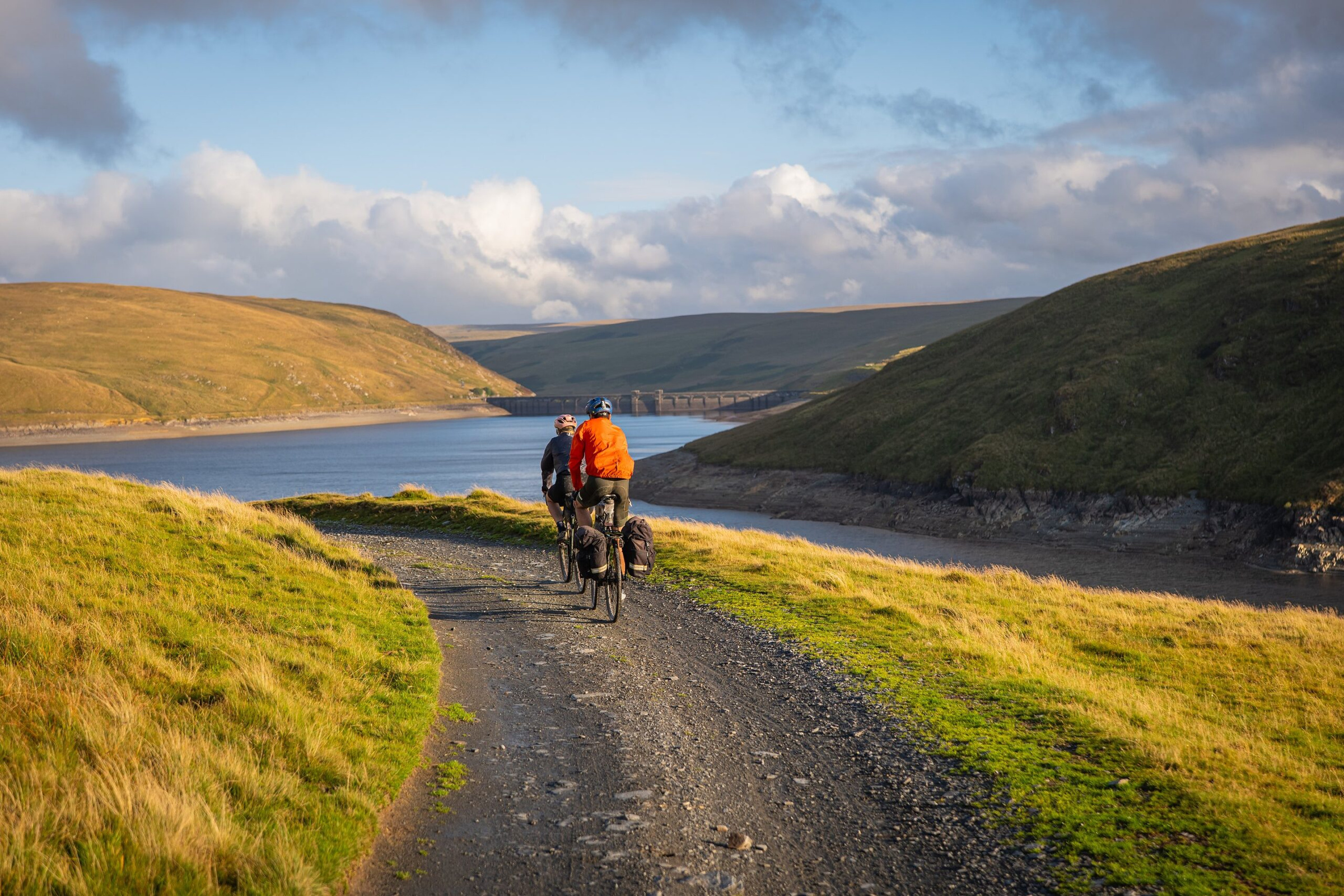 Two riders on a gravel track beside Claerwen reservoir in evening light